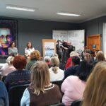 Dwie kobiety stoją przed zgromadzonymi w sali ludźmi. W tle wystawa posterowa. Kamerzysta kieruje obiektyw na przemawiającą osobę.